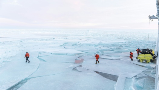 Forskere i oransje overlevelsesdrakter arbeider på isflak i et arktisk landskap, med et gult kjøretøy og et skip ved kanten av bildet.