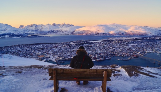 En person sitter på en benk og ser ut over Tromsø by, omgitt av snødekte fjell og hav i solnedgang.