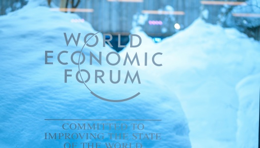 Logoen til World Economic Forum på et vindu, med store snøfonner og et bygg i bakgrunnen. Teksten under logoen lyder «Committed to improving the state of the world».