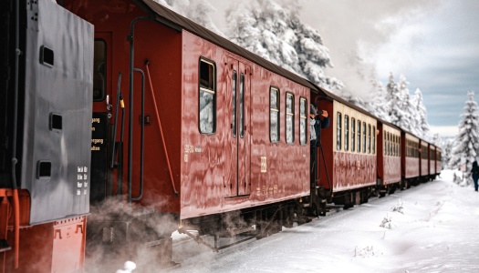Et damptog med røde og gule vogner kjører gjennom et snødekt landskap omgitt av trær. Dampen stiger fra toget mens en person tar bilder ut av vinduet.