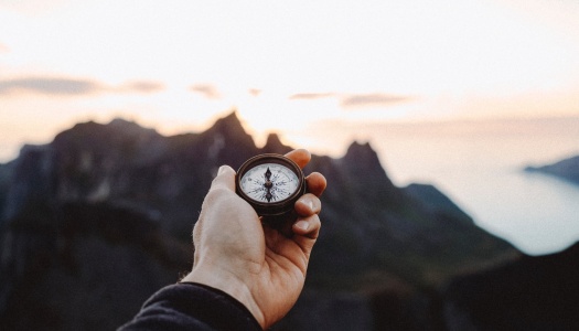 En hånd holder et kompass i forgrunnen, med uklare fjell og en lysende horisont i bakgrunnen ved solnedgang.