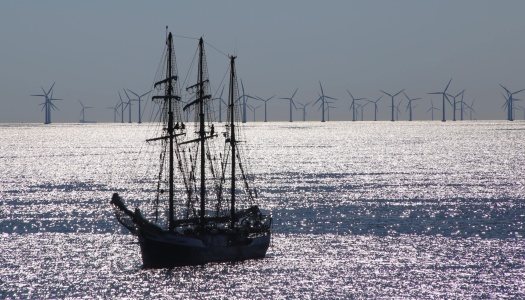 Et seilskip ligger på glitrende hav med en rekke vindmøller til havs i bakgrunnen, som illustrerer kontrasten og sameksistensen mellom tradisjonell sjøfart og moderne fornybar energi.