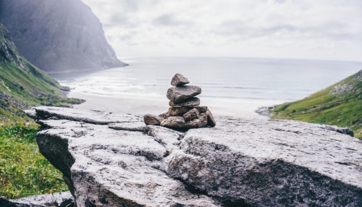 En liten varde av steiner står på en fjellhylle med utsikt mot havet og en strand omgitt av grønne fjellsider.