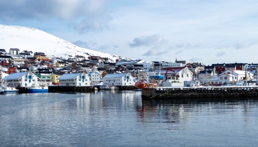 Kystby med fargerike hus og fiskebåter ved kai, omgitt av snødekte fjell og rolig hav i nordnorsk vinterlandskap.