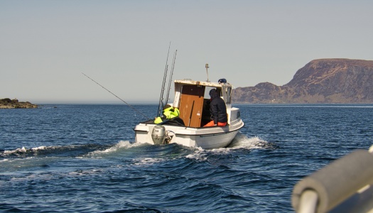 En liten fiskebåt med to personer i arbeidsklær kjører utover på åpent hav med fiskestenger om bord, med fjell og kystlandskap i bakgrunnen.