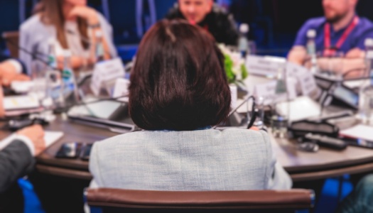 Deltakere i diskusjon rundt et rundt konferansebord, sett bakfra fra en av deltakerne.