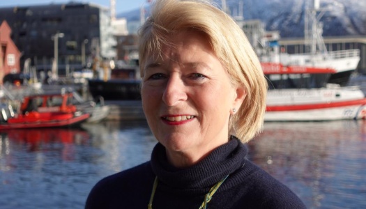 Kvinne med blondt hår smiler foran en havn med båter og bygninger i bakgrunnen på en solrik dag.