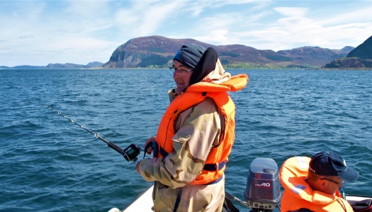 To personer fisker fra en båt på åpent hav omgitt av fjell og blå himmel, iført redningsvester.