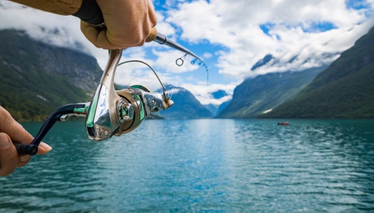 En person fisker med en snell i klart vann omgitt av fjell og solskinn, trolig i en norsk fjord.