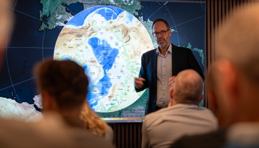 Foredragsholder peker og forklarer foran et stort kart over Arktis på en skjerm, med publikum sittende i forgrunnen.