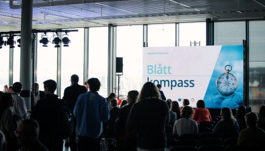 Publikum i en sal. En skjerm med et bilde som sier "Blått kompass"
