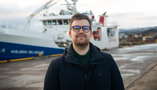 En person med kort hår og briller står utendørs på en kai. I bakgrunnen ligger et stort skip fortøyd ved havn, med kran og overbygg synlig. Personen har på seg en mørk jakke, og det er overskyet vær.