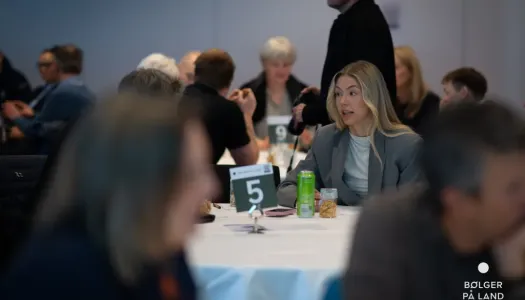 Kvinne sitter ved bord nummer 5 og deltar i samtale under et arrangement, med andre deltakere rundt bordet og personer i bakgrunnen. Logoen «Bølger på land» vises nederst til høyre.