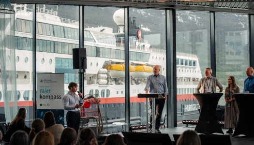 Paneldebatt på konferansen Blått kompass, med flere deltakere på scene foran store vinduer med et Hurtigruten-skip i bakgrunnen.