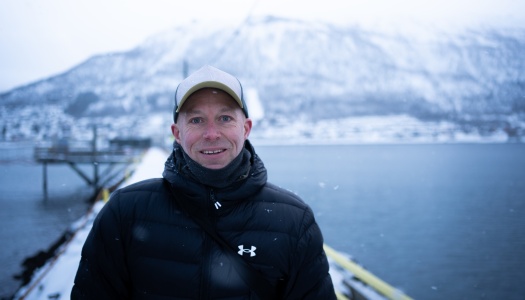 En smilende mann i vinterklær står på en brygge ved vannet, med snødekte fjell i bakgrunnen og lett snøfall rundt seg.