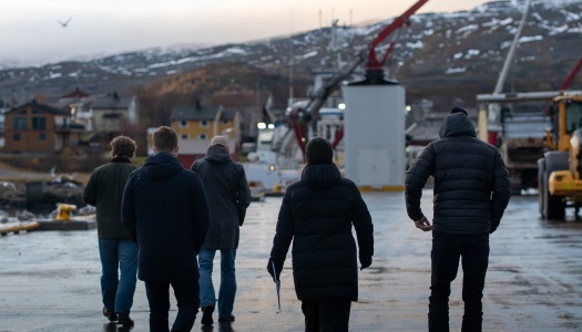 Fem personer går langs en kai i et lite kystsamfunn med snødekte fjell i bakgrunnen og industrielt utstyr rundt seg.