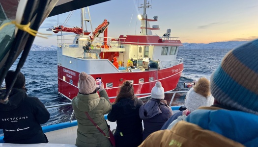 Turister i varme klær står på dekk av en båt og tar bilder av en rød fiskebåt på havet i arktisk landskap ved solnedgang.