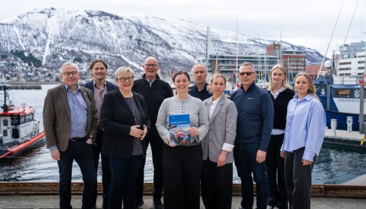 En gruppe på ti personer står samlet ved en havn med båter og snødekte fjell i bakgrunnen. En kvinne i midten holder en rapport, og alle vender mot kameraet.