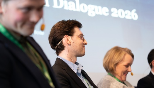 Tre personer med hodesett sitter på en scene under et arrangement, med teksten «Dialogue 2026» synlig i bakgrunnen.
