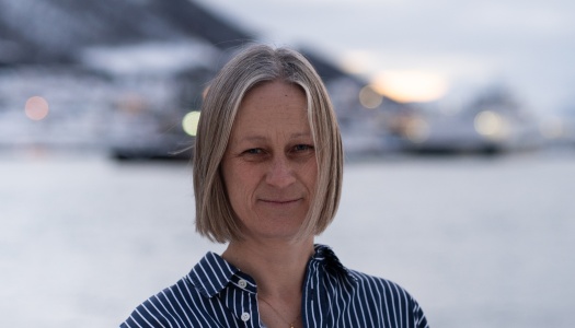 Portrett av en kvinne med kort, lyst hår som står utendørs ved vannet, med snødekte fjell og bebyggelse i bakgrunnen.