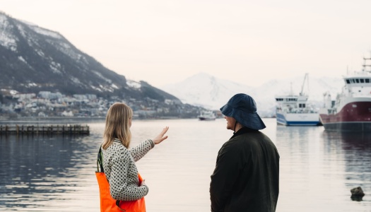 To personer står ved en havn og ser ut over vannet mot båter og snødekte fjell.