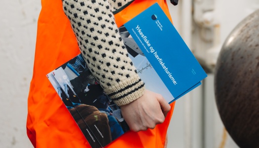 Person i oransje arbeidsklær holder en blå brosjyre med tittelen ‘Yrkesfiske og havbrukslære’, stående utendørs