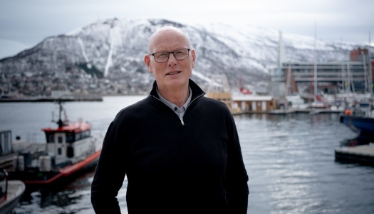 Mann med briller står ved en havn, med båter og snødekte fjell i bakgrunnen.