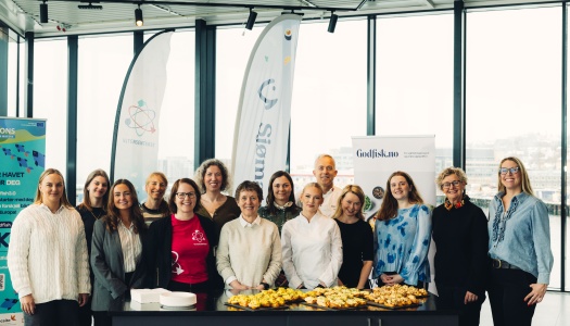 En gruppe på rundt 14 personer står samlet bak et bord med småretter i et lyst lokale, smilende mot kamera, med bannere i bakgrunnen.