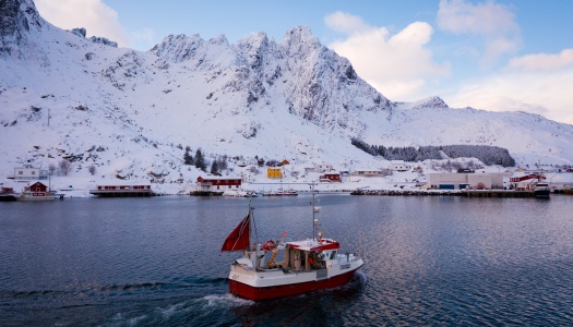Fiskebåt på vei inn mot et snødekt kystsamfunn omgitt av bratte fjell i nordnorsk vinterlandskap.