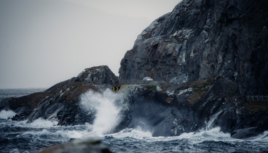 Bølger slår mot en bratt og værutsatt kystvei langs havet. En bil kjører på den smale veien mellom mørke klipper og opprørt sjø.