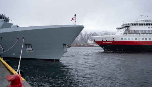 Grått marinefartøy og Hurtigruteskipet Otto Sverdrup ligger ved kai i Tromsø, med snødekte fjell i bakgrunnen.