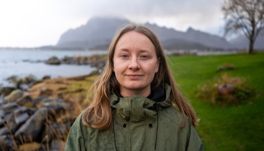 Therese Hugstmyr Woie står ved en steinete kystlinje med havet og fjell i bakgrunnen. Hun har på seg en grønn jakke og ser mot kameraet.