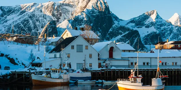 Små fiskebåter fortøyd ved en snødekt havn med hvite trehus og høye fjell i bakgrunnen, opplyst av den lave vintersola i Lofoten, Norge.