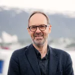 Portrett av mann i dressjakke med briller, smilende foran hav og fjell i bakgrunnen.