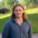 Portrett av en mann med langt hår og blå skjorte, stående utendørs foran en grønn plen