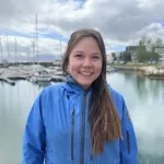 ortrett av en smilende kvinne i blå jakke, stående foran en marina med båter og vann.