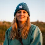 Kvinne med mørkeblondt hår ikledd lue og fleecegenser ser rett på kameraet med et lite smil. 