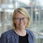 Kvinne med skulderlangt blondt hår ser rett inn i kamera med et smil. Hun har på briller og blå dressjakke. 