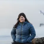 Portrett av en kvinne med mørkt hår i blå jakke, som smiler utendørs ved sjøen.