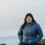 Portrett av en kvinne med mørkt hår i blå jakke, som smiler utendørs ved sjøen.