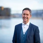 ortrett av en smilende mann i dress som står utendørs ved vannet, med by og bro i bakgrunnen.