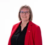 Person med kort hår og briller smiler mot kameraet, iført rød blazer og sort skjorte foran en lys bakgrunn.