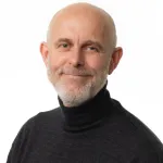 Person med kort skjegg og mørk høyhalset genser smiler rolig mot kameraet foran en lys bakgrunn.