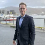 «En mann i dress står utendørs ved en havn, med båter og fjell i bakgrunnen, og ser mot kameraet.