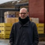 Person med briller og mørk jakke står utendørs foran stablede plastkasser og industribygg.
