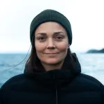 Person iført mørk jakke og grønn lue står utendørs foran et hav og fjell i bakgrunnen på en overskyet dag.