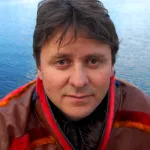 Person i tradisjonell samisk kofte ser rett mot kamera, med vann i bakgrunnen.