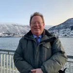 En mann står utendørs foran et vinterlandskap med snødekte fjell, vann og en by i bakgrunnen. Han har på seg briller, en vinterjakke og smiler mot kameraet.
