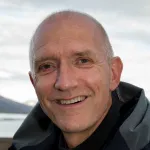 Smilende person i friluftsjakke står utendørs med fjell og vann i bakgrunnen.