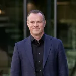 Person i mørk dress og svart skjorte står utendørs foran en glassfasade, og ser rolig mot kameraet.